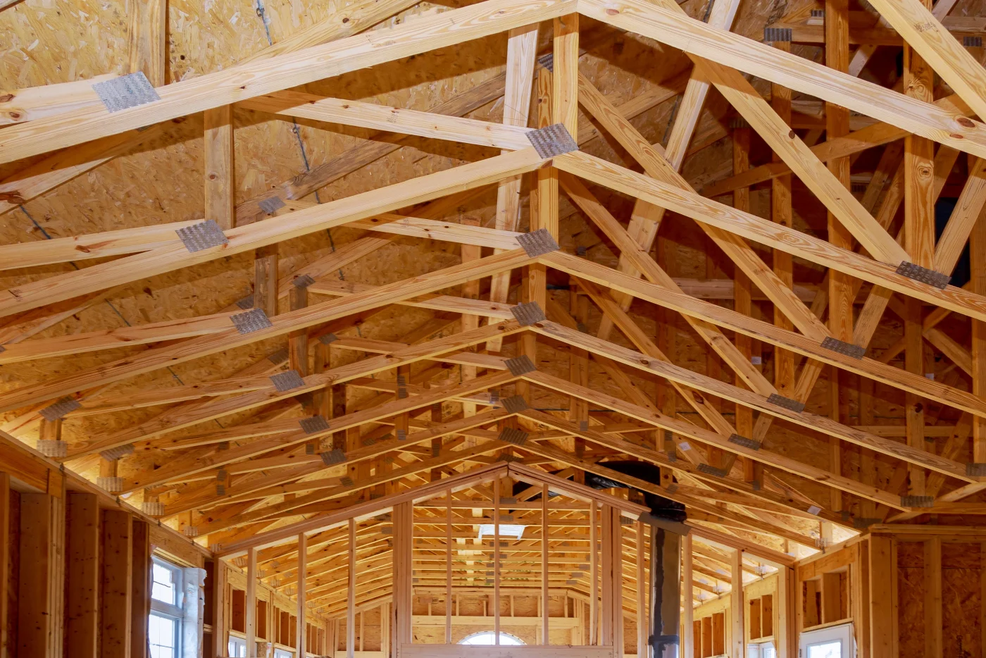 The inside of a long, narrow building with exposed wooden framing on the walls and the ceiling. The ceiling has multiple visible trusses, and there are flat wooden boards above them acting as the roof’s base. Light can be seen coming in through multiple windows toward the back of the pictured area.