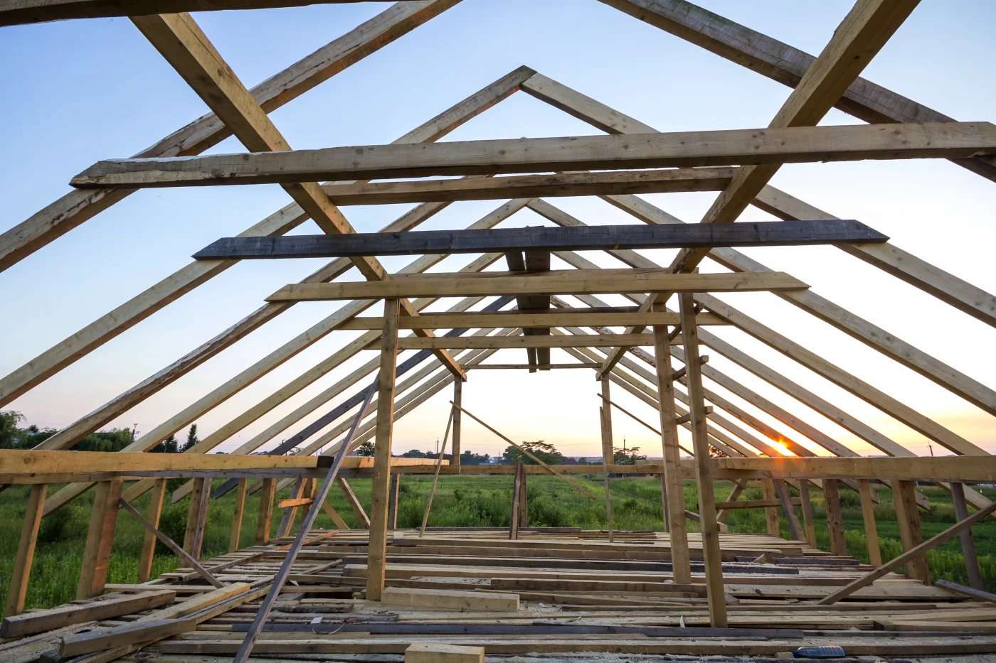Elite Barndominium Truss In Warrensburg, MO | North Country Truss A photo taken from the floor of an attic, looking up at the truss system that has been set to support the ceiling and roof. Beyond the roof and upper level frame, there is a large, green field and a sunset visible.