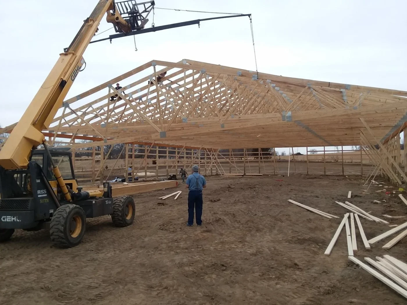 A photo of a large crane lifting a wooden truss into place during the construction of a low, wide agricultural building. A man wearing a blue shirt and suspenders is standing at the center of the image and watching the process. There are wooden boards on the ground on the right side of the picture.