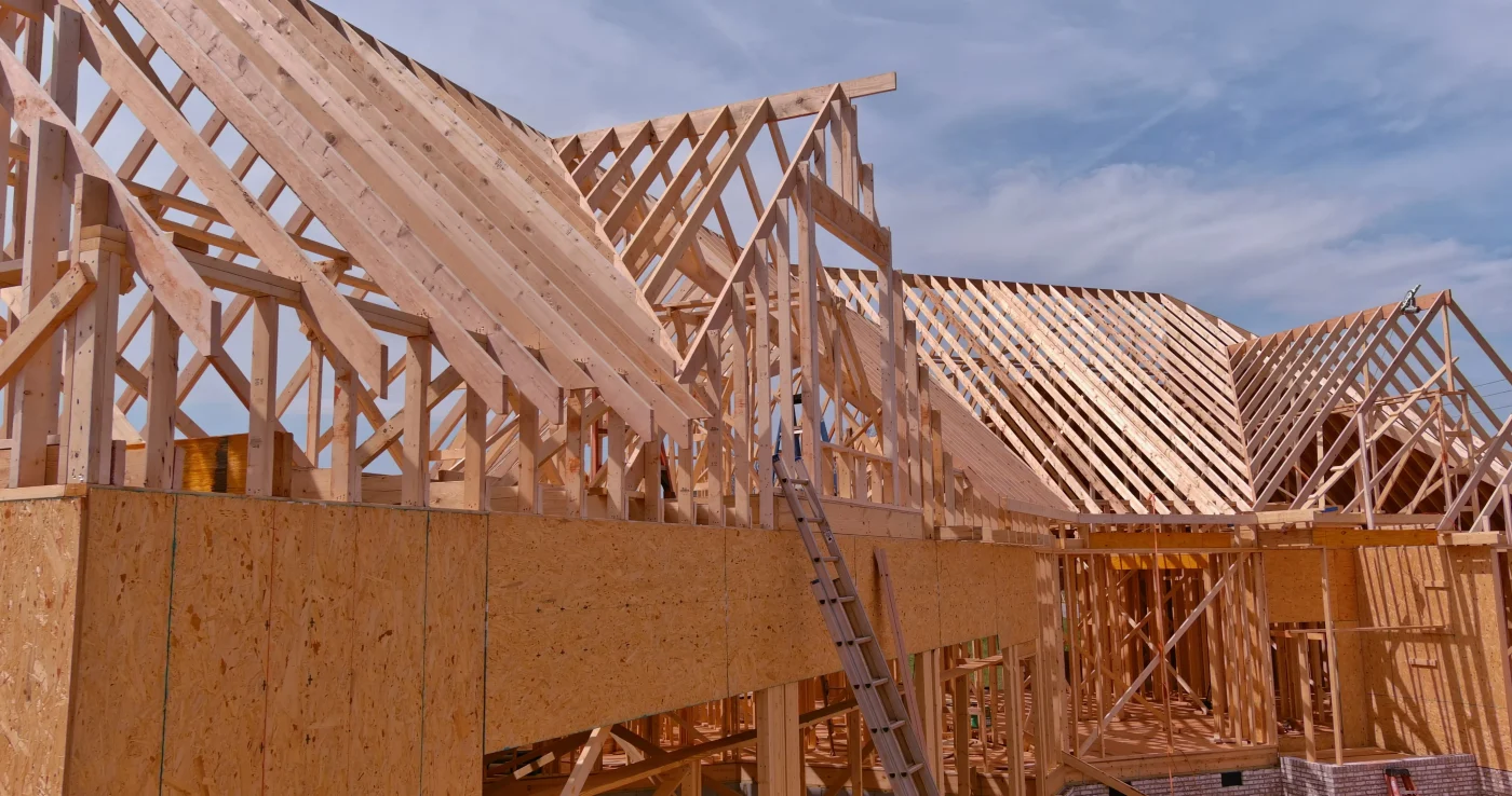 A large, sprawling house under construction. Some of the plywood wall framing is up, and above that, the roof and remaining walls are framed out. A ladder can be seen leaning up against one of the front walls.
