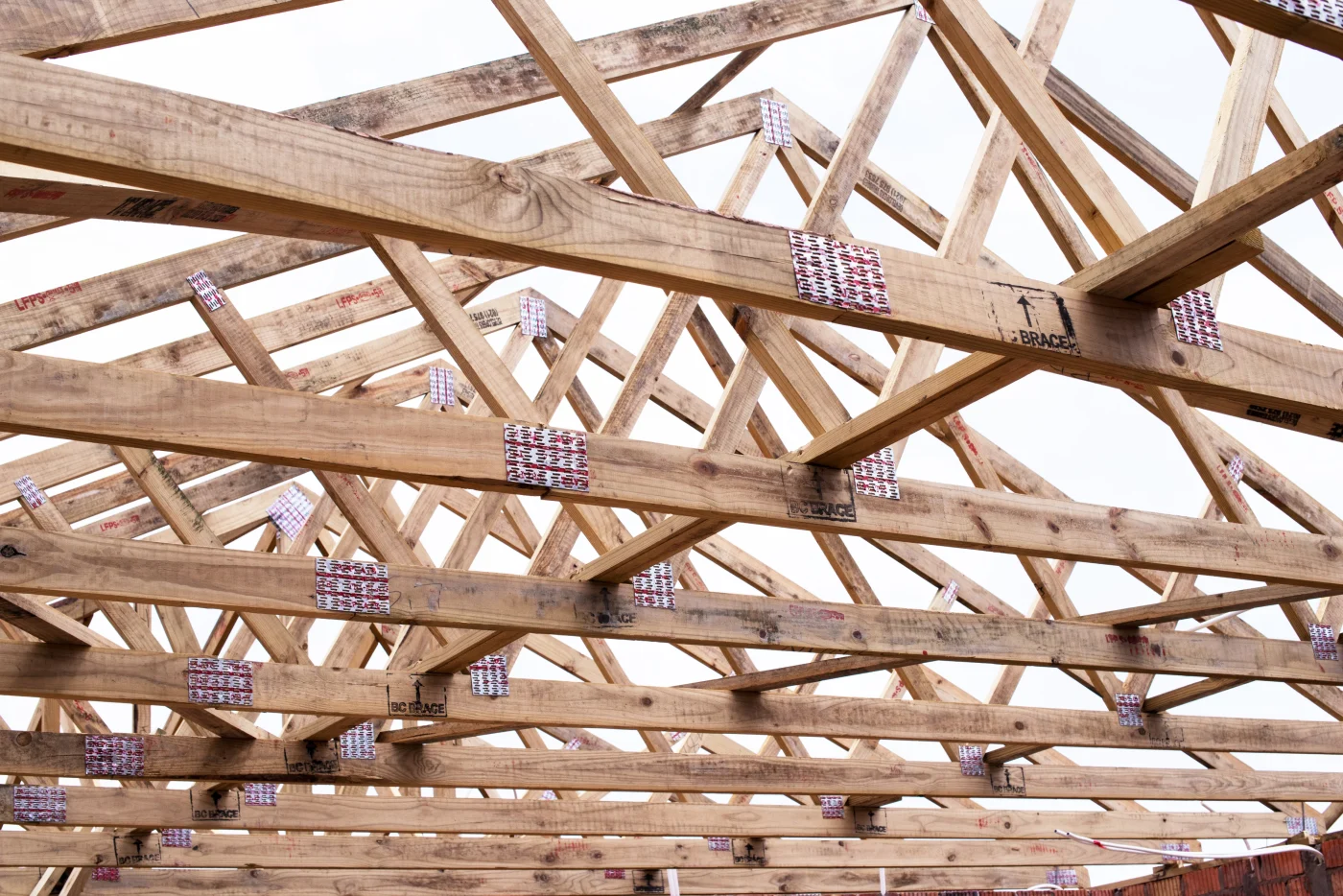 A close-up showing a roof’s wooden framing and truss arrangement with open sky above it. The truss boards all have visible metal connector plates at the joints.