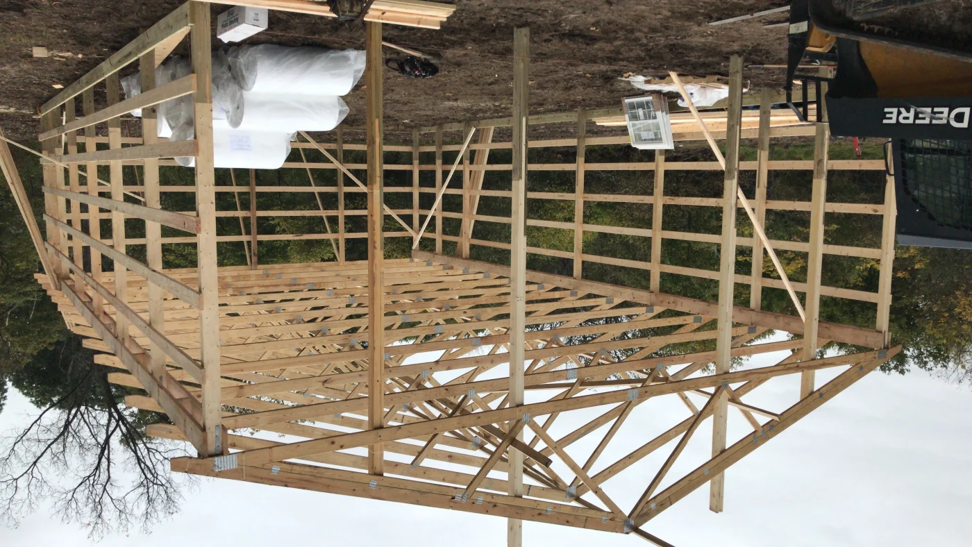 A close-up of the wooden framing for a small, wide barn. The roof is partially in place, with an evenly spaced truss system, and assorted building materials can be seen on the ground inside the framed-out structure.
