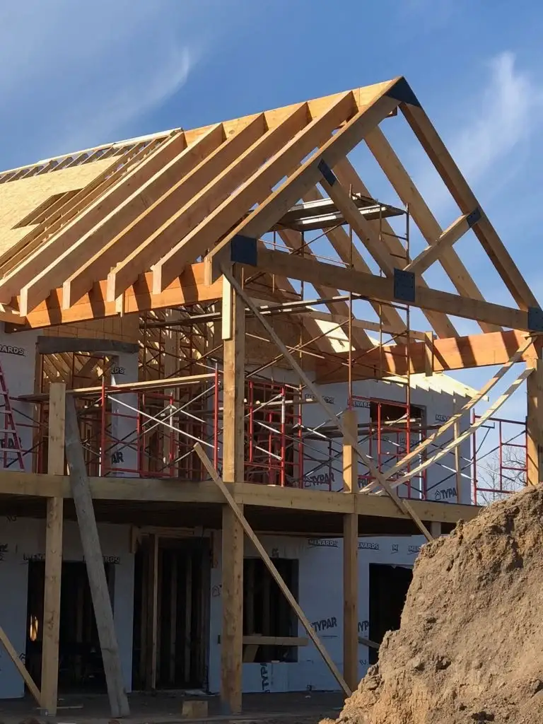 Enchanting Barndominium Truss In Warrensburg, MO | North Country Truss A photo taken of a home in the middle of construction showing off the truss system in the roof’s frame above an extended upper level patio. The frame is wood, and there is red scaffolding beneath it to act as support while it’s under construction. The exterior of the house has a white wrap on it.