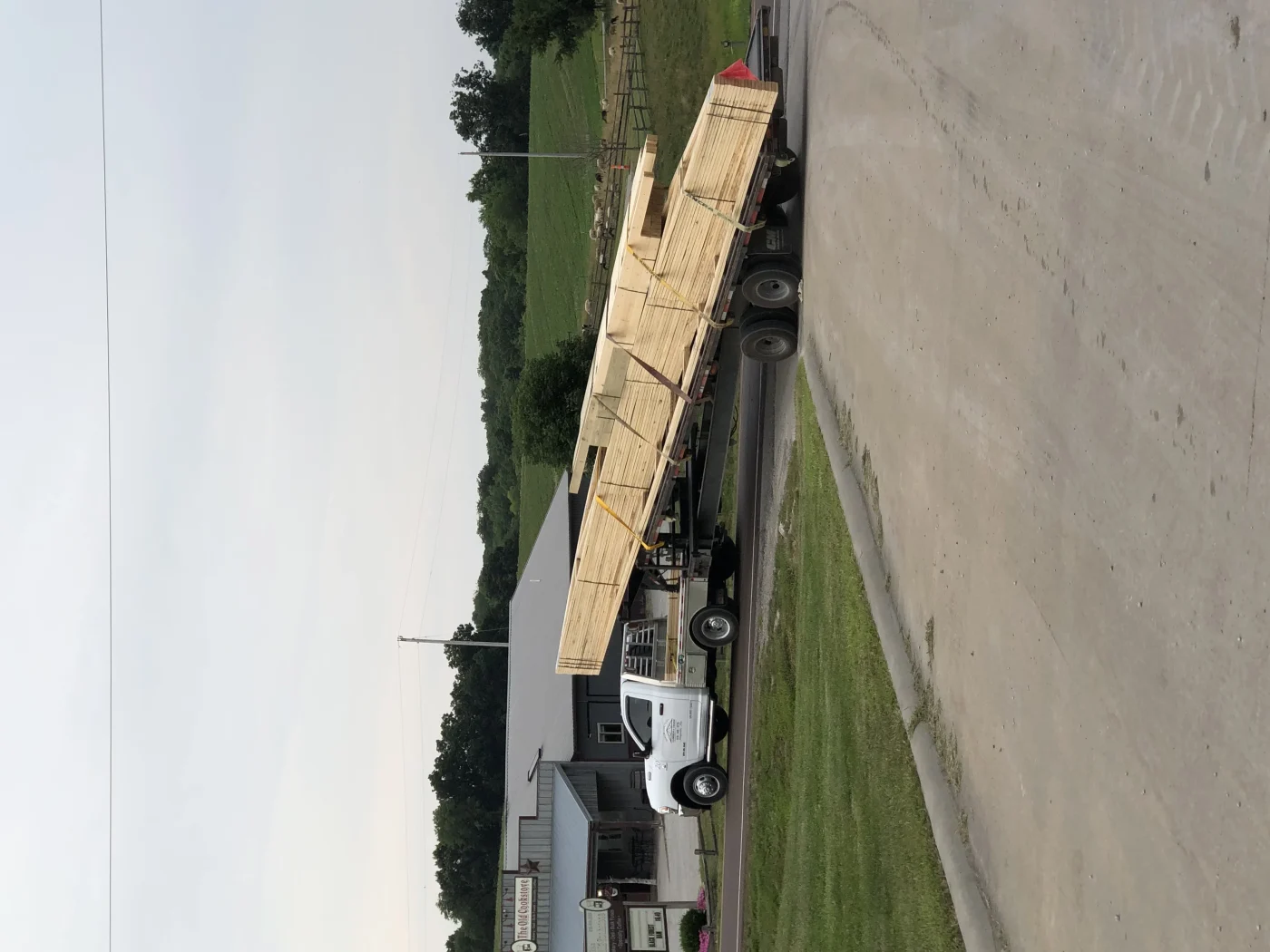 A photo showing a flat-bed truck with a load of wooden trusses turning onto a road. The truck’s cab is white, and there are multiple trusses stacked and tied down for transport.