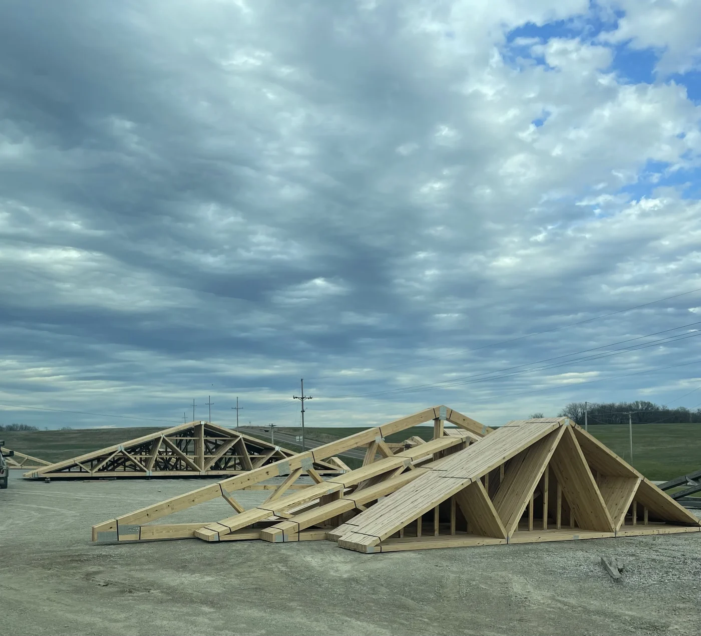 A photo showing multiple pre-built trusses standing up and grouped together. There are multiple sides and designs for support visible, and in the back, there are multiple larger trusses visible. They’re arranged on a concrete slab in front of a large, open field.